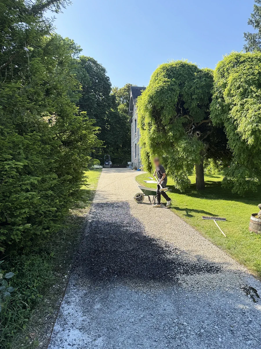 Compagnon répandant le liant sur une allée bordée d'ifs, manoir en arrière-plan