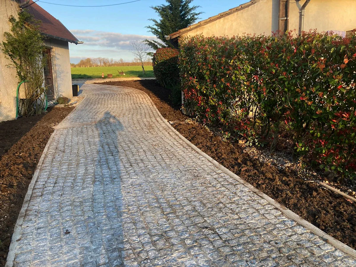 Allée en pavés au tracé courbe entre une maison et une haie de photinias, terre travaillée de part et d'autre