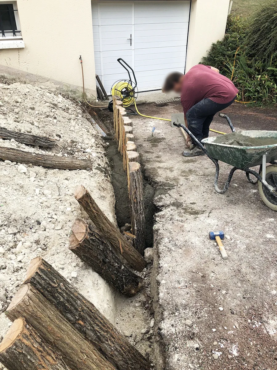 Compagnon scellant une bordure en rondins de bois dans une tranchée le long d'un garage