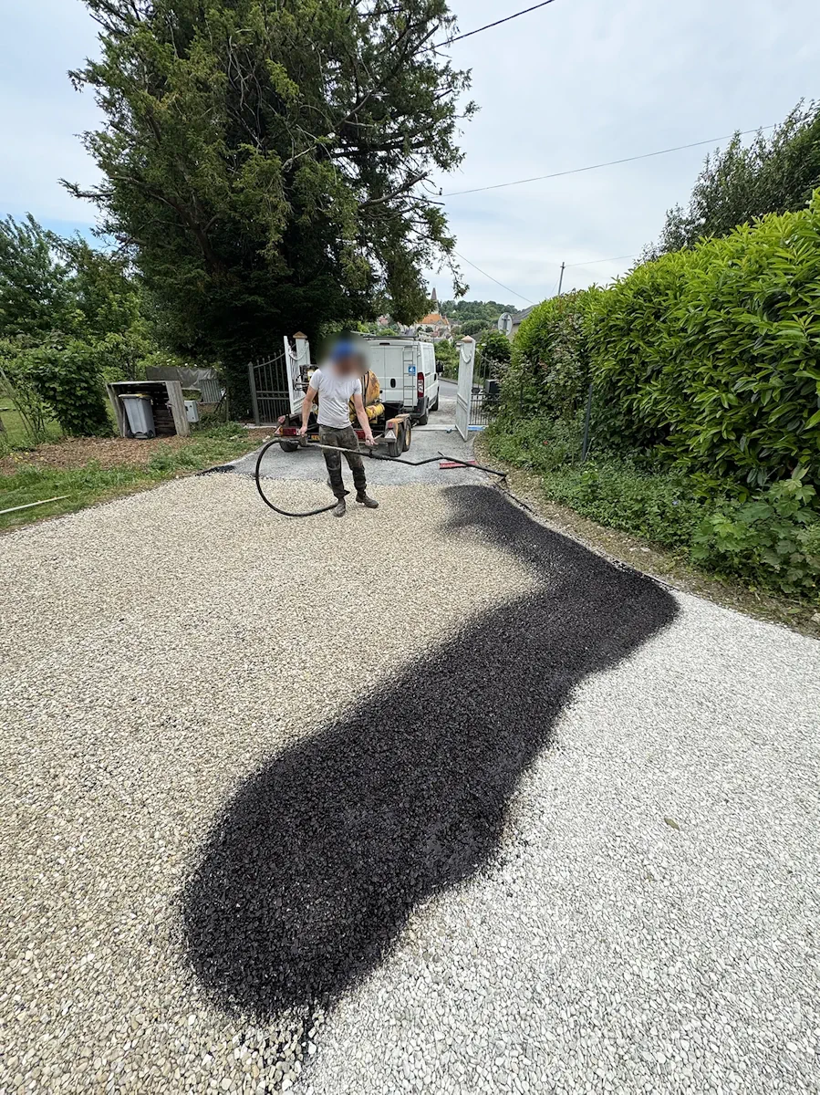 Compagnon appliquant le liant sur une cour en gravier, portail et haie en arrière-plan