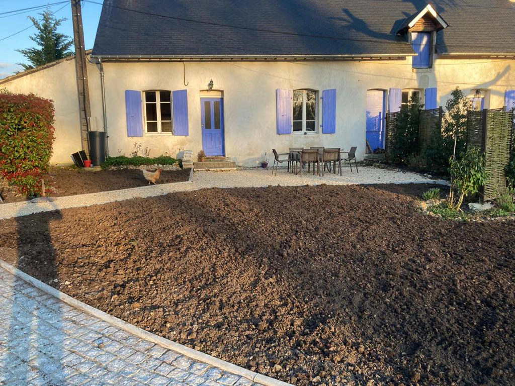 Cour d'une maison aux volets bleus, terre nivelée en attente d'engazonnement, bordure pavée posée