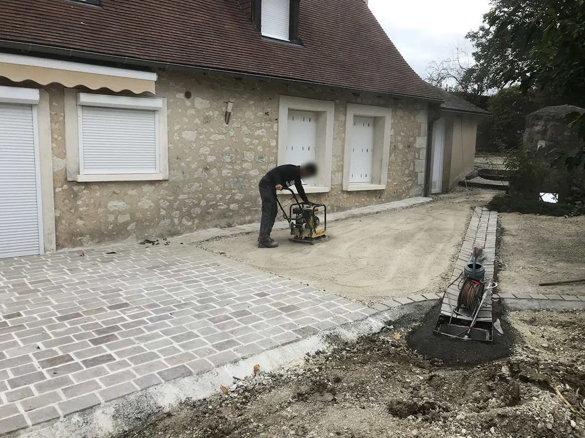 Compagnon compactant le lit de sable d'une cour, pavage déjà posé sur une partie