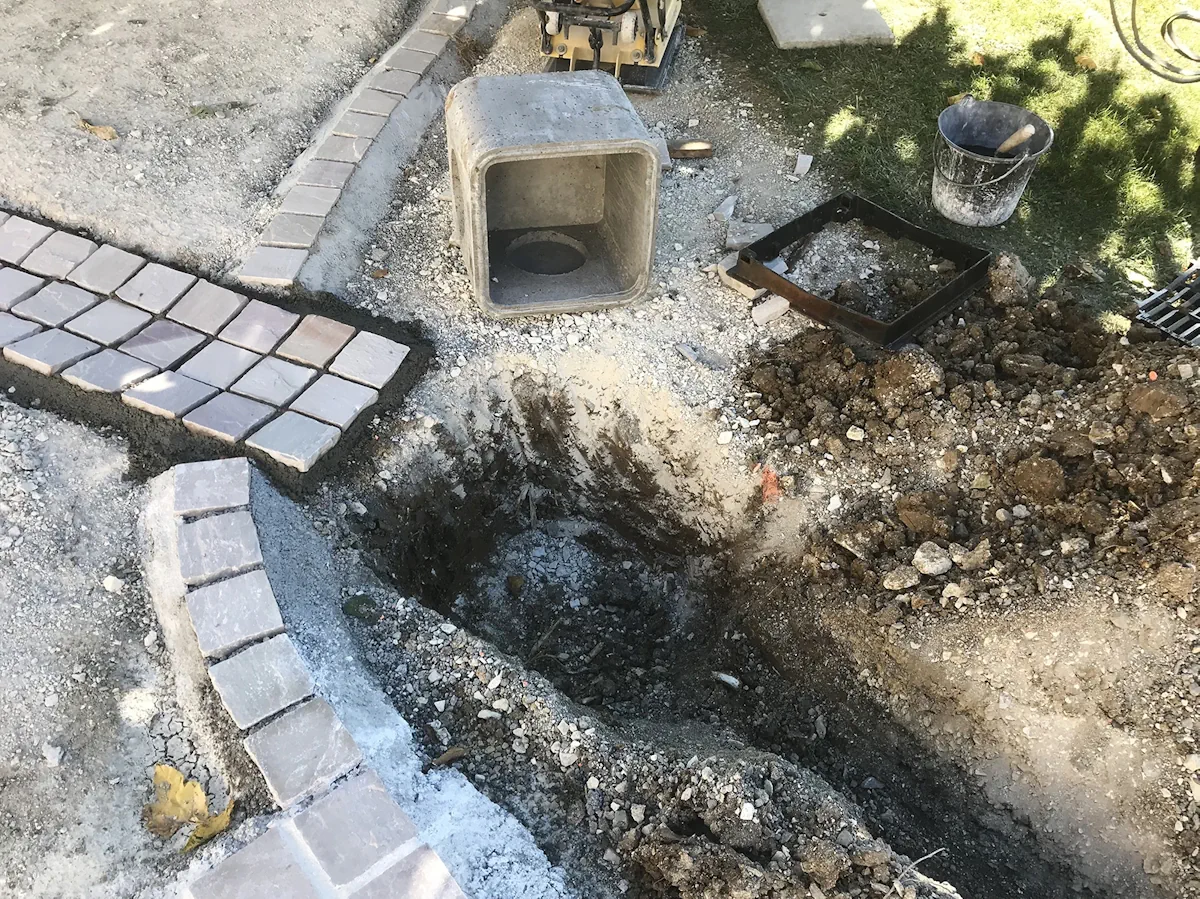 Regard béton posé au bord d'une allée pavée, tranchée ouverte pour le raccordement