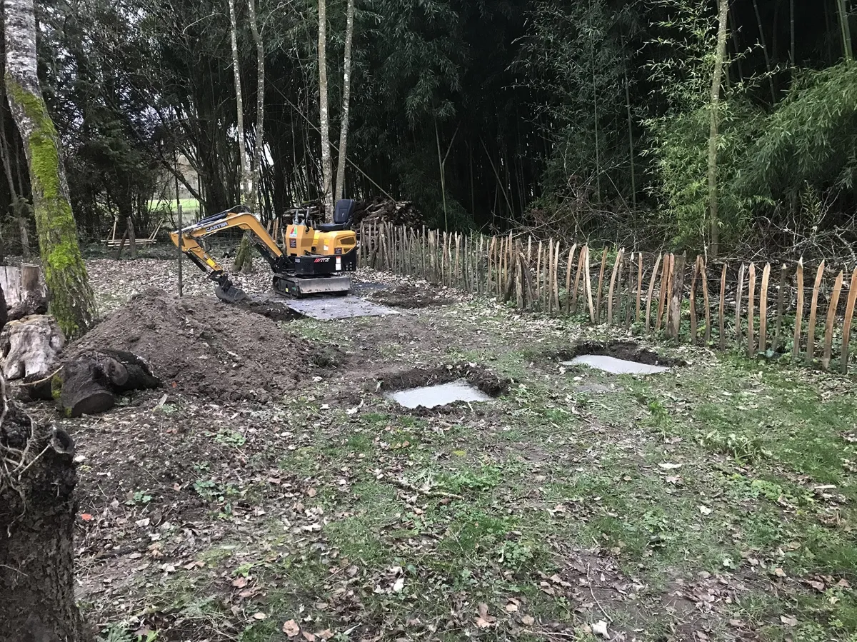 Mini-pelle terrassant au pied d'une ganivelle de châtaignier, plots béton en attente