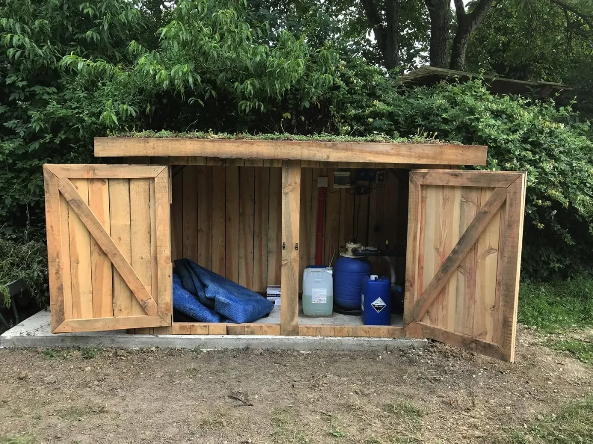 Local technique de piscine en bardage bois à toiture végétalisée, portes ouvertes sur le groupe de filtration