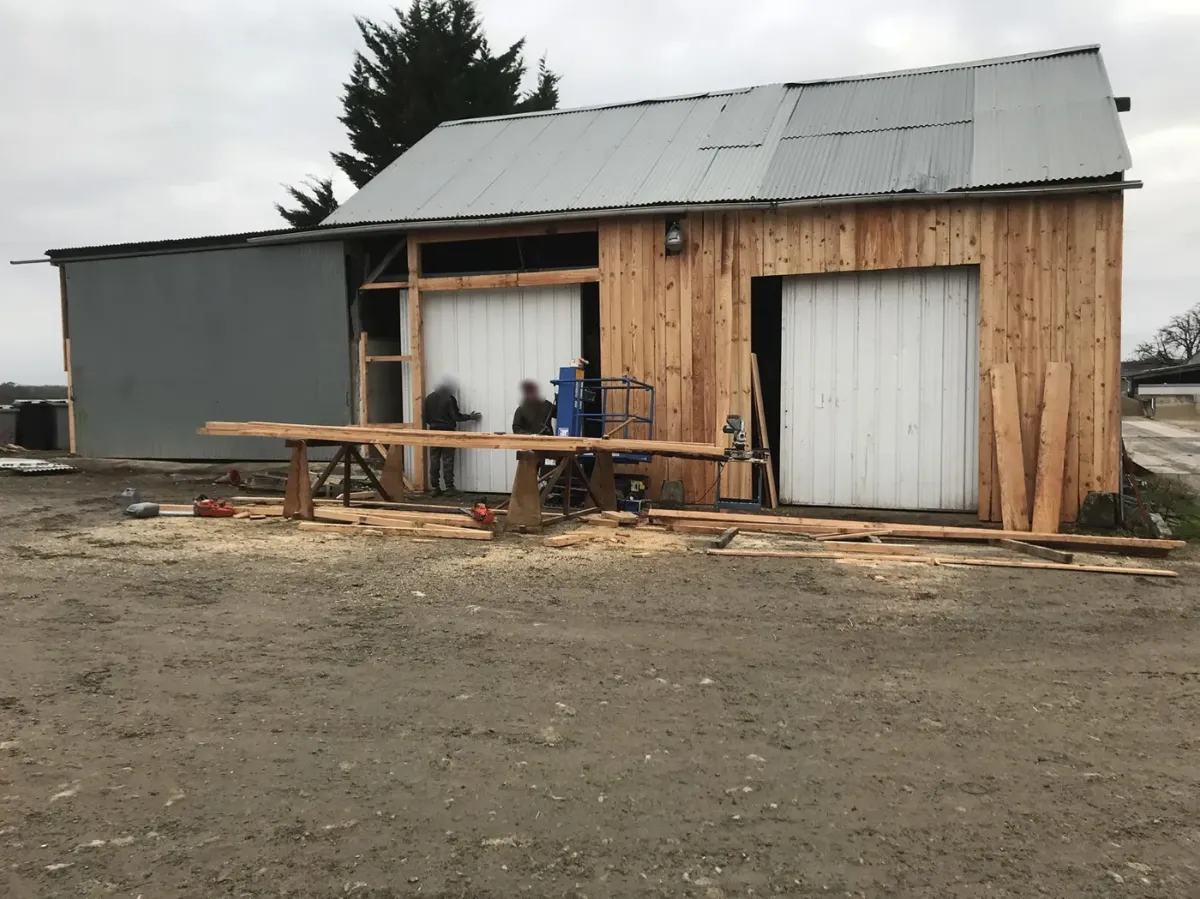 Deux compagnons posant le bardage bois d'un hangar agricole, tréteaux et scie sur l'aire de chantier