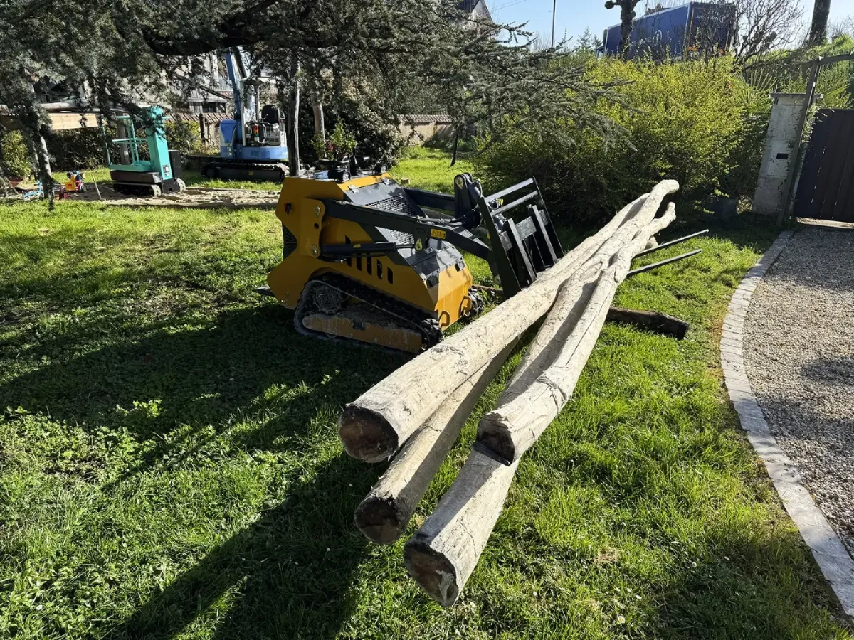 Chargeuse compacte à fourches devant trois longs poteaux de bois écorcés posés sur l'herbe