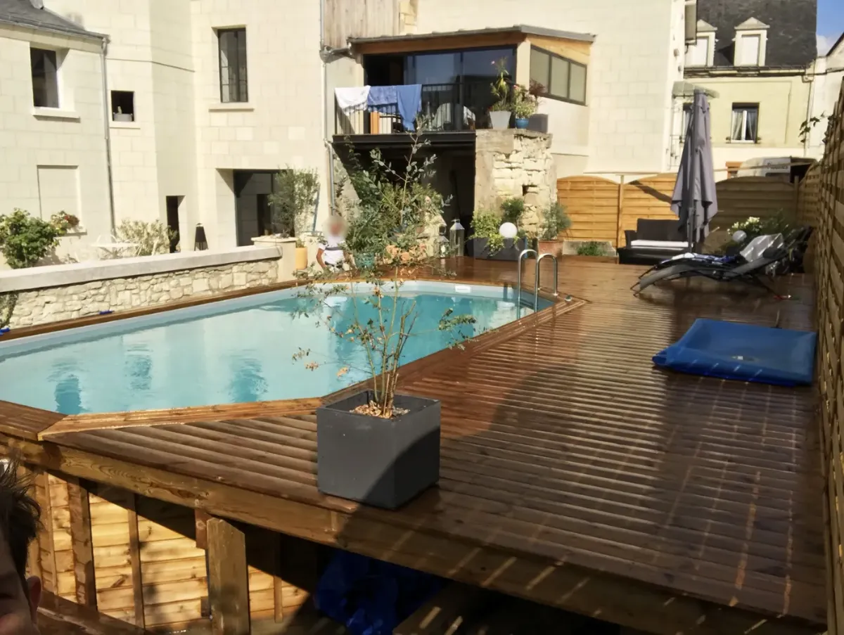 Terrasse en bois entourant une piscine dans une cour close de bâtiments en tuffeau