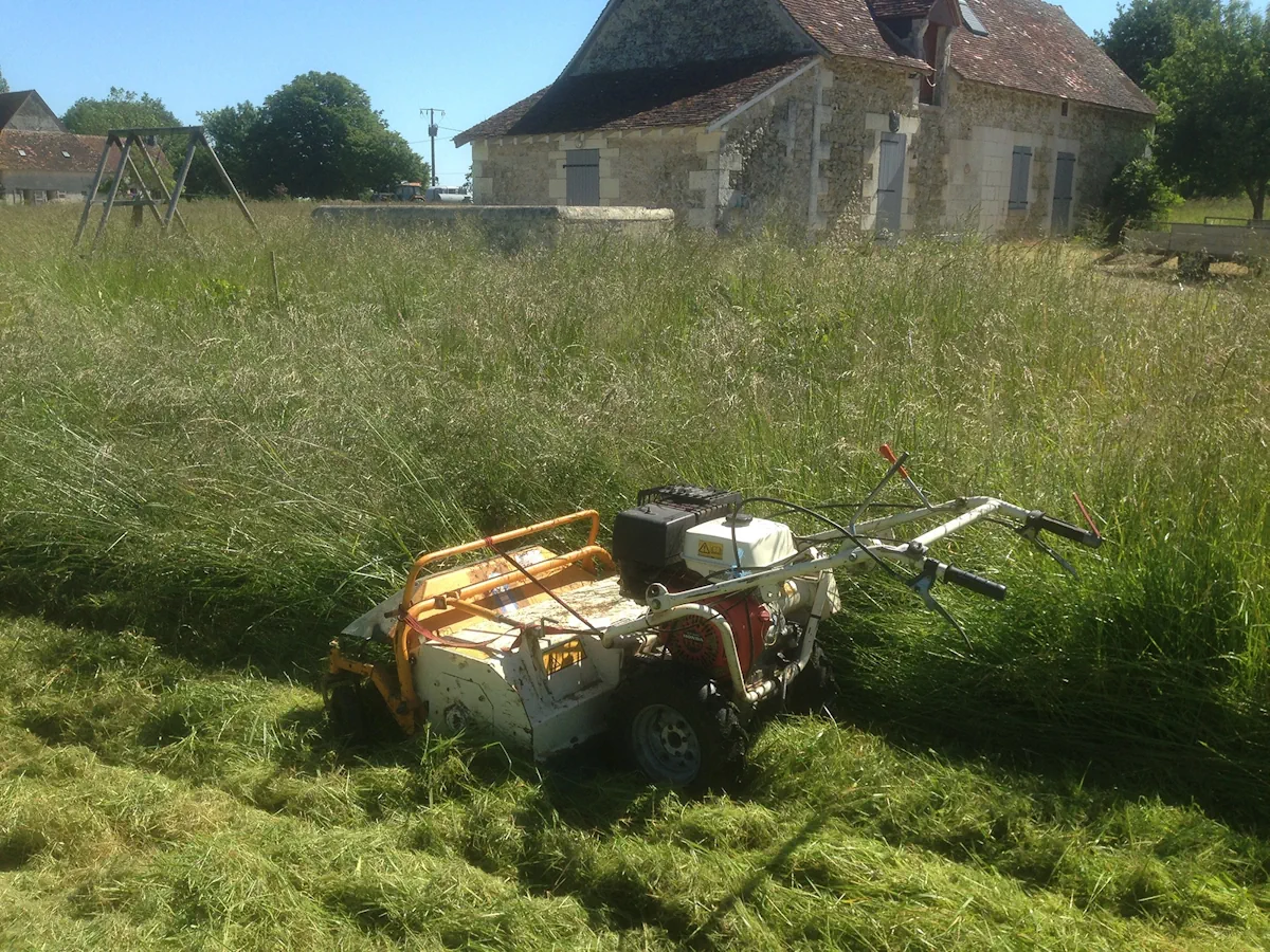 Débroussailleuse autotractée arrêtée dans l'herbe haute devant une longère et son portique