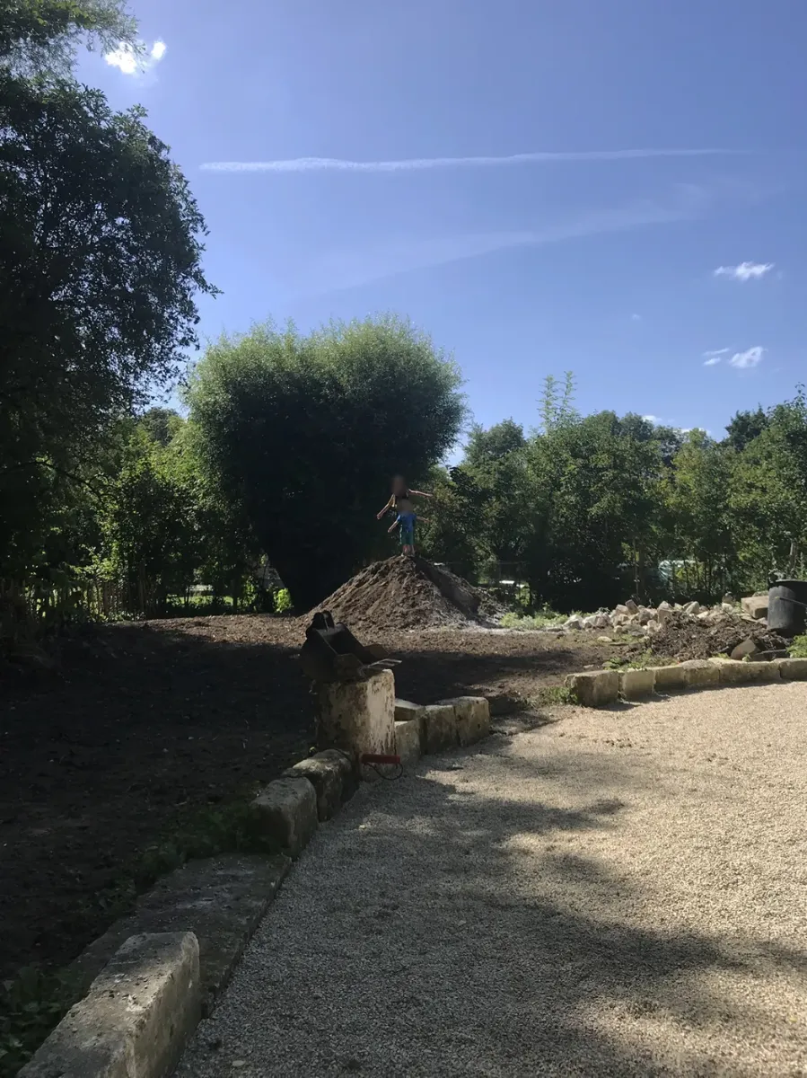 Terrain en cours de terrassement, godet de pelle posé au sol et gros tas de terre au centre, deux enfants juchés au sommet