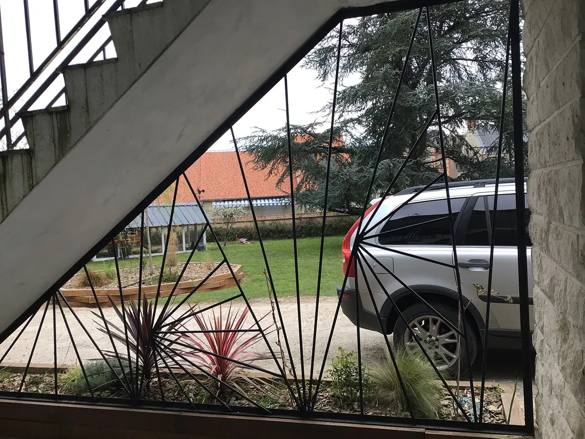 Grille d'acier à motif rayonnant vue depuis l'intérieur, ouvrant sur la cour et le jardin