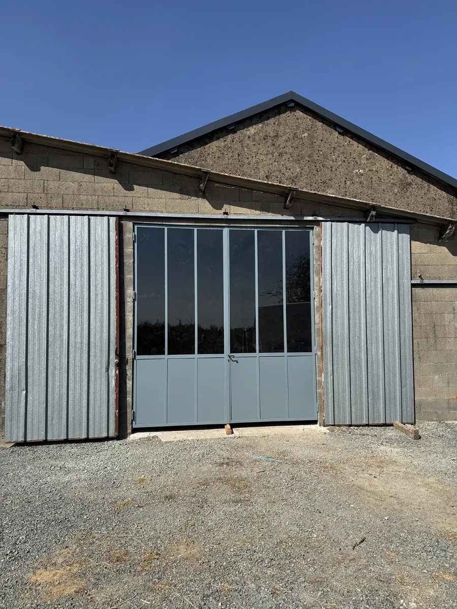 Verrière d'atelier en acier gris installée dans le pignon d'un hangar, entre deux portes de tôle