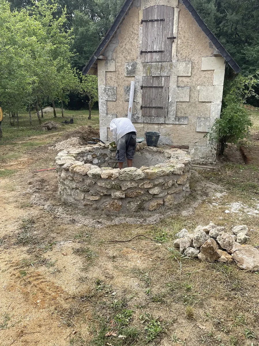 Compagnon montant à la main la margelle circulaire en pierre d'un puits, devant une maisonnette