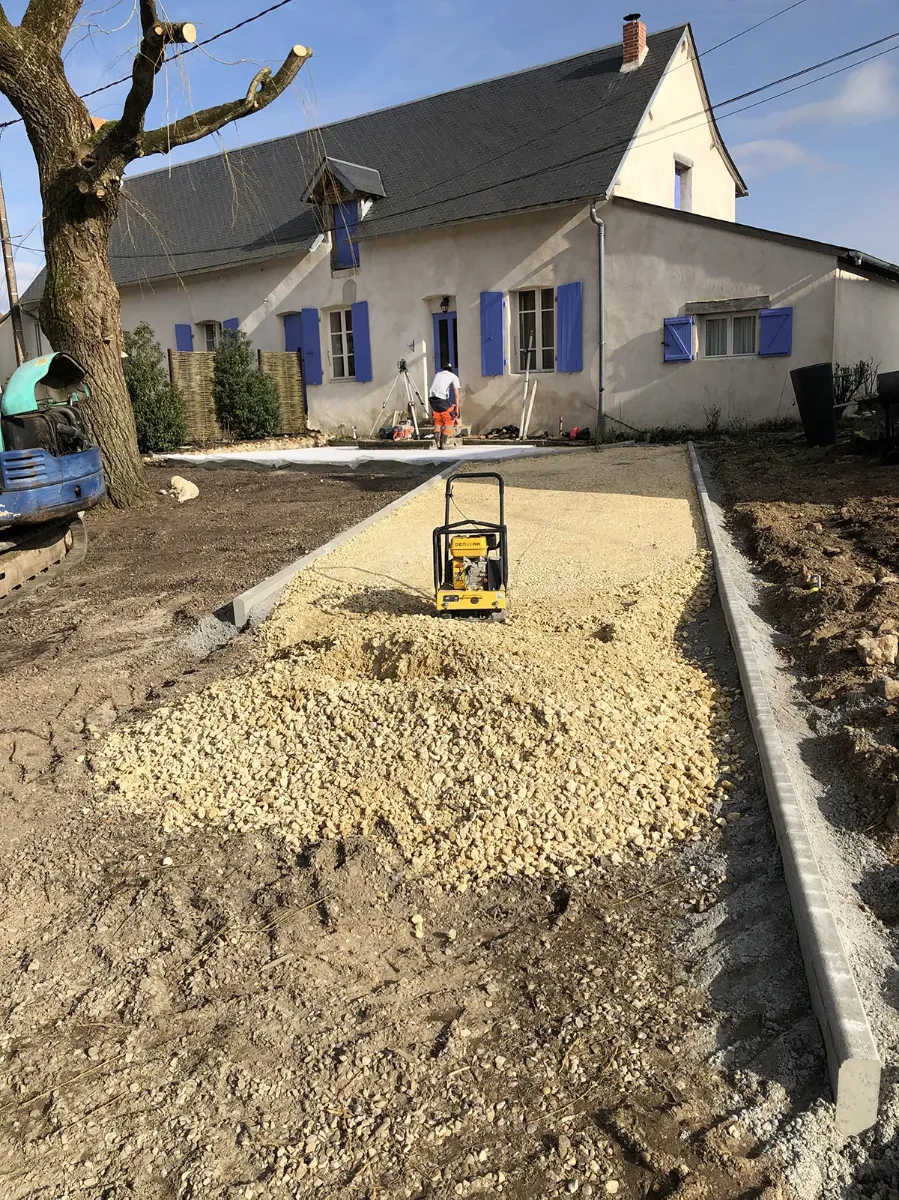 Plaque vibrante compactant le tout-venant d'une allée entre deux bordures béton, devant une longère