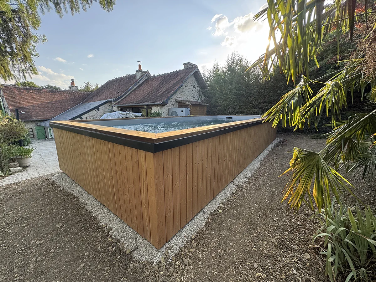 Piscine hors-sol bardée de bois et coiffée de noir, devant une longère de pierre, au soleil couchant