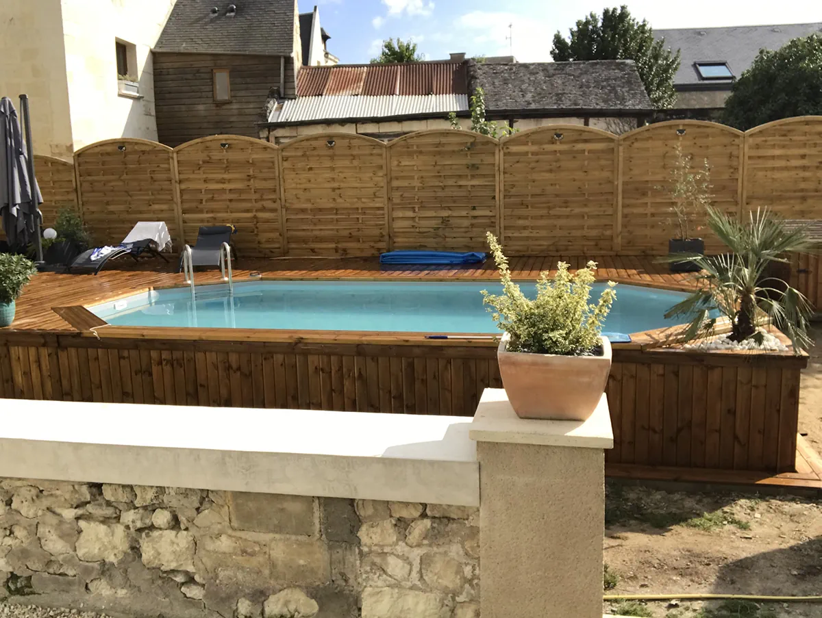 Piscine bois et sa terrasse vues par-dessus un muret de pierre, palissades cintrées au fond