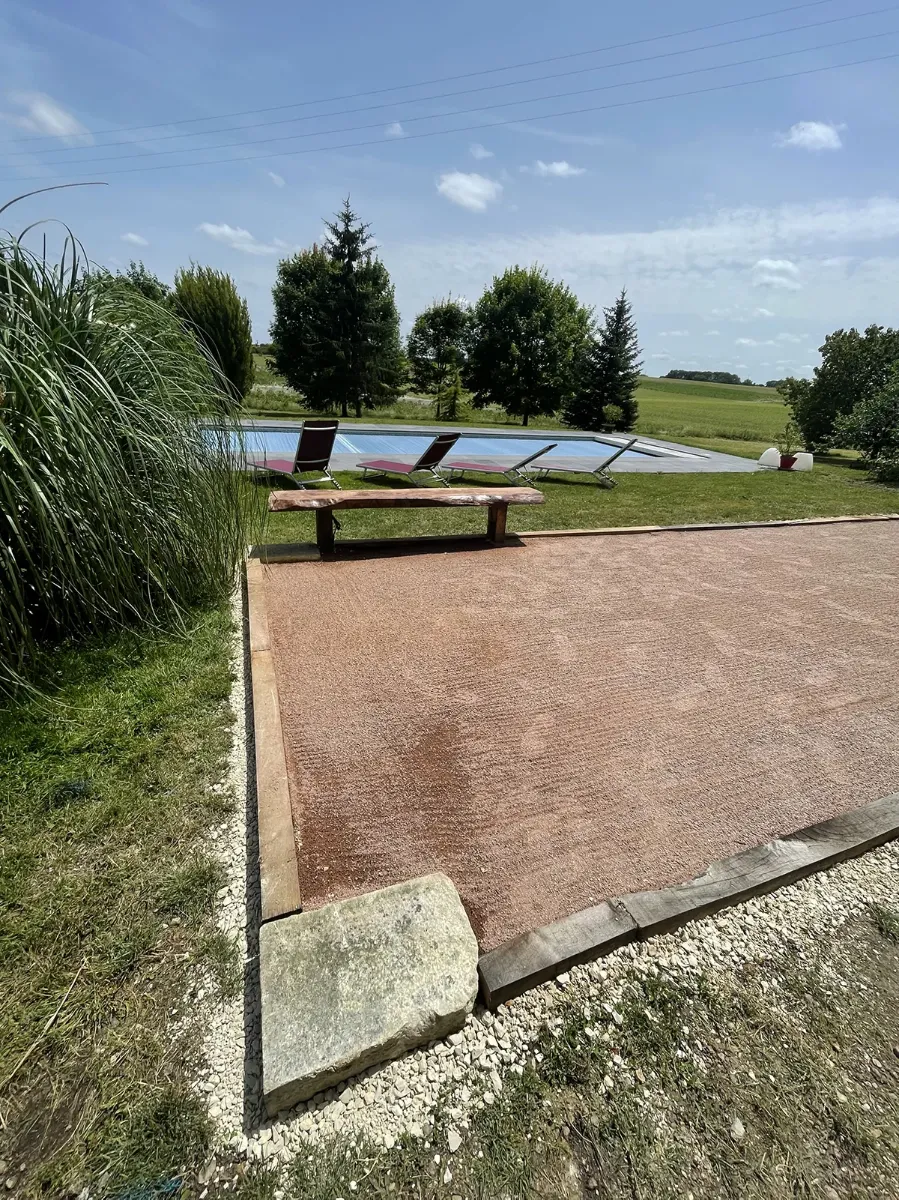 Terrain de boules en mâchefer rose bordé de madriers de chêne, banc de bois et piscine ouverte sur les champs