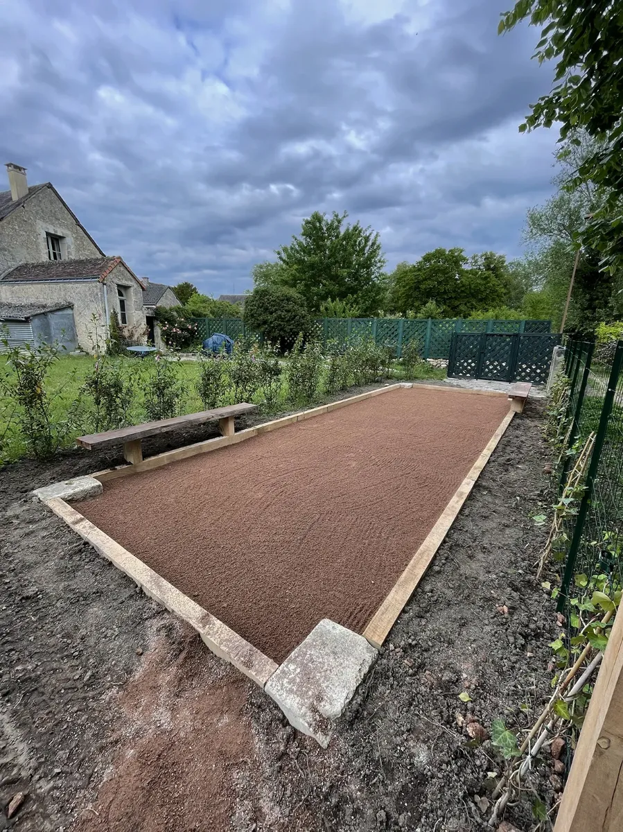 Terrain de boules neuf bordé de madriers et de blocs de pierre, devant une longère, sous un ciel d'orage
