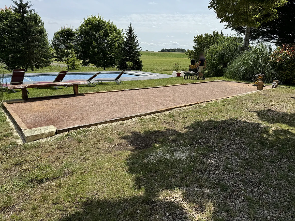 Terrain de boules en mâchefer vu depuis son extrémité, banc de bois et piscine sur la gauche