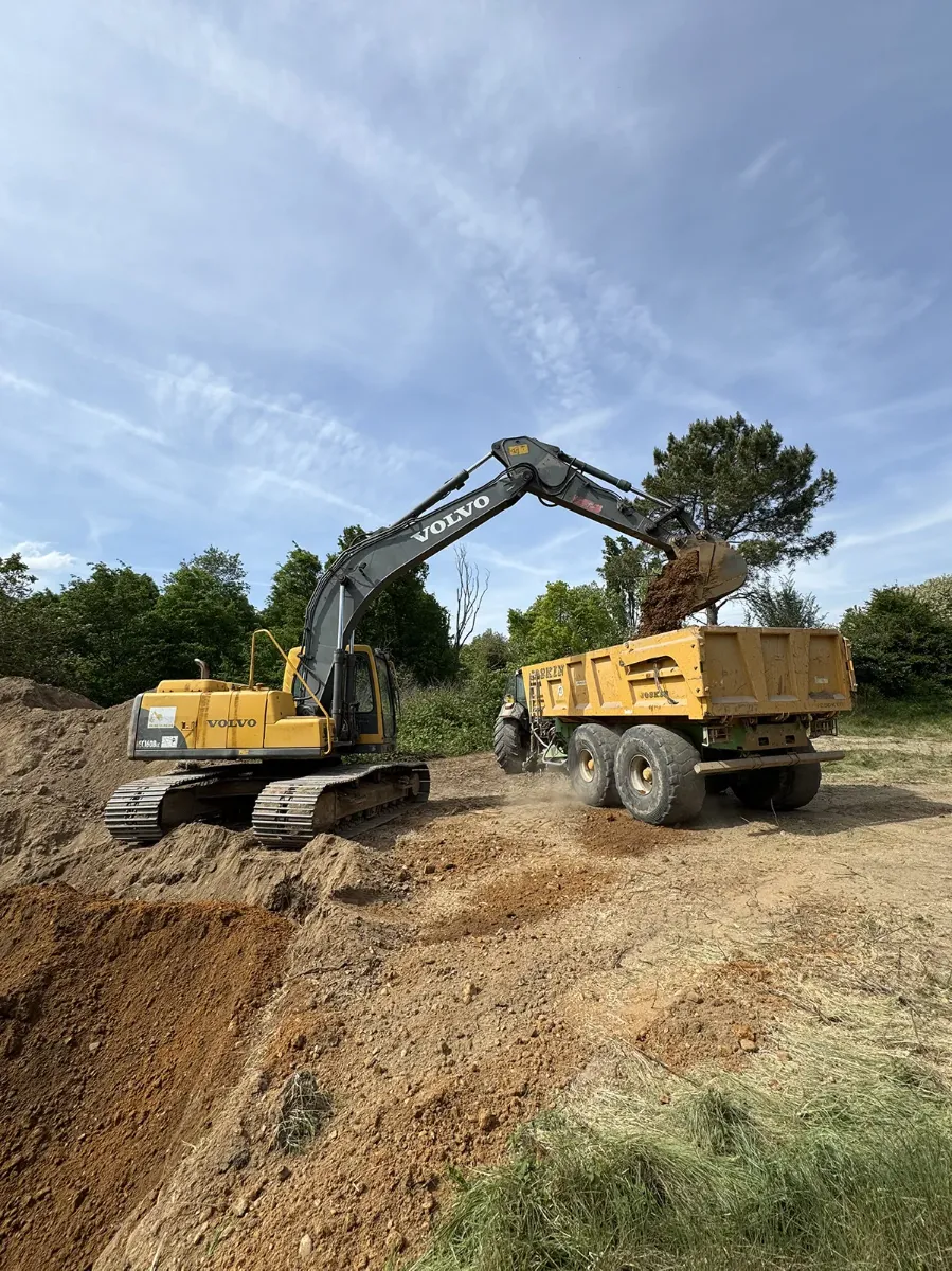 Pelle Volvo chargeant une benne agricole en terre, chantier de terrassement en bordure de bois