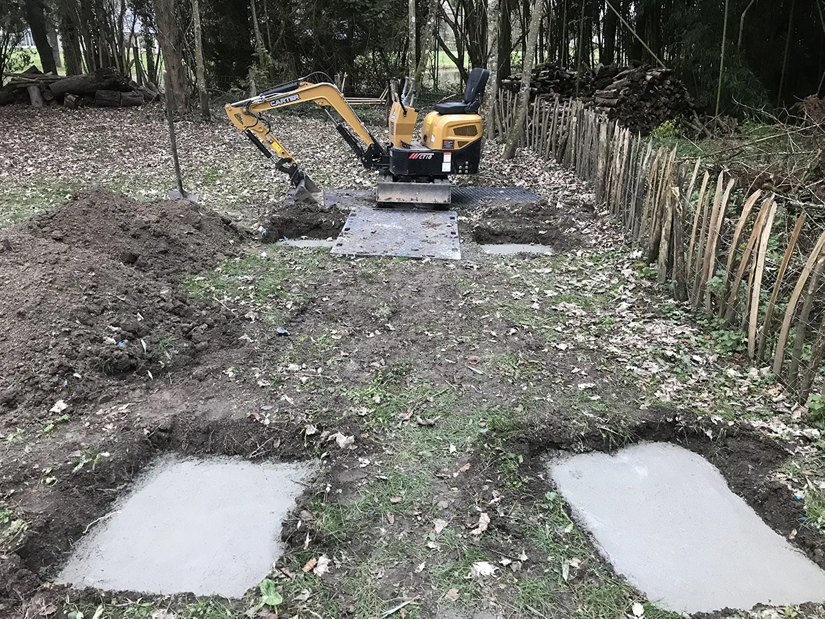 Quatre plots de béton coulés en sous-bois pour recevoir une construction, mini-pelle sur ses plaques de roulage