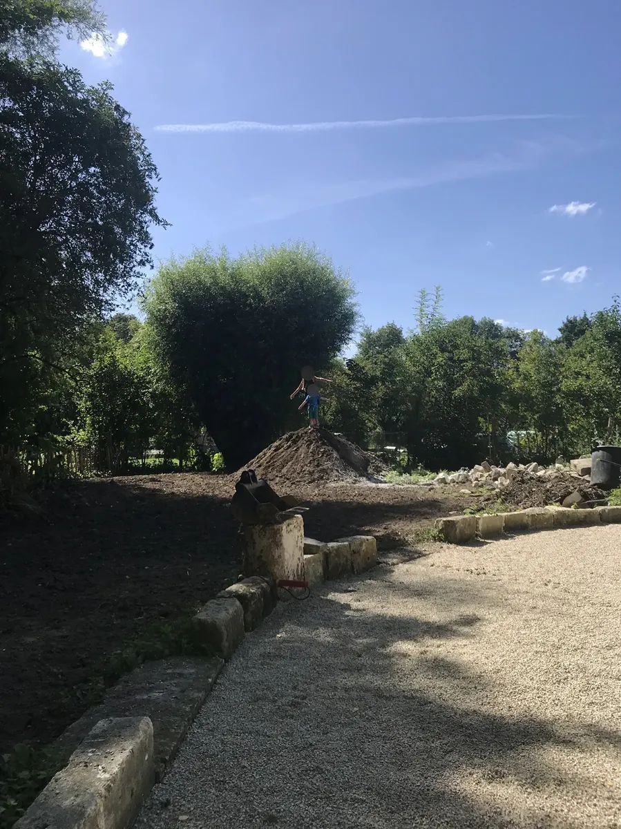 Tas de terre et godet déposé en bordure d'une cour de gravier, un enfant juché au sommet, en plein été