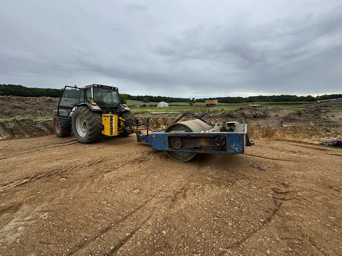 Tracteur attelé à un rouleau compacteur sur une plateforme de terrassement, sous un ciel couvert