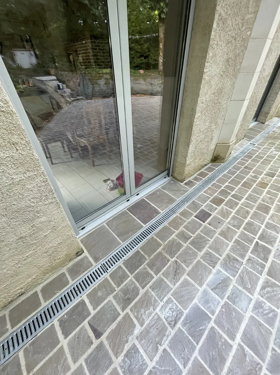 Caniveau de façade posé au ras d'une baie vitrée, le long d'un calepinage de pavés de pierre