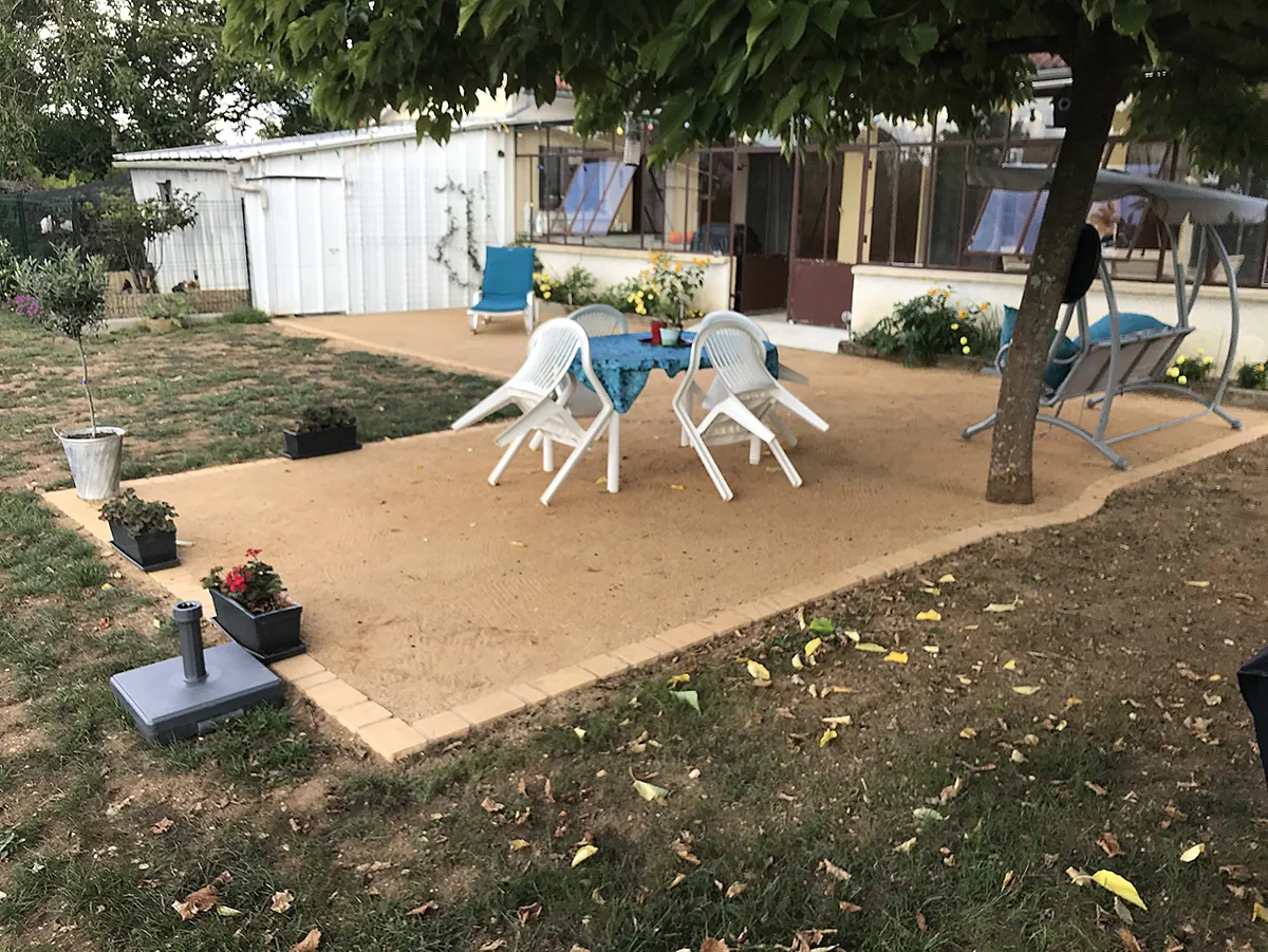Terrasse de sable stabilisé bordée de pavés, aménagée sous un grand arbre avec table et fauteuils
