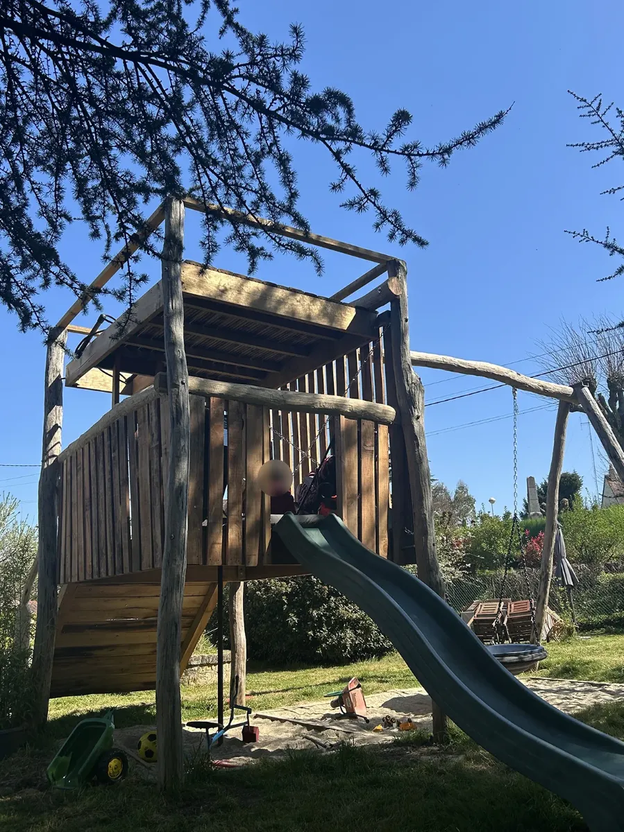 Cabane de jeux sur pilotis en rondins, avec plancher, palissade et toboggan, au-dessus d'un bac à sable