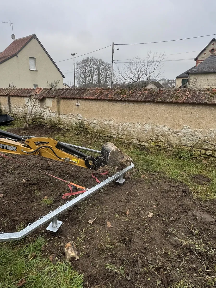 Châssis galvanisé de trampoline sanglé au bras d'une mini-pelle, en cours de positionnement dans un jardin