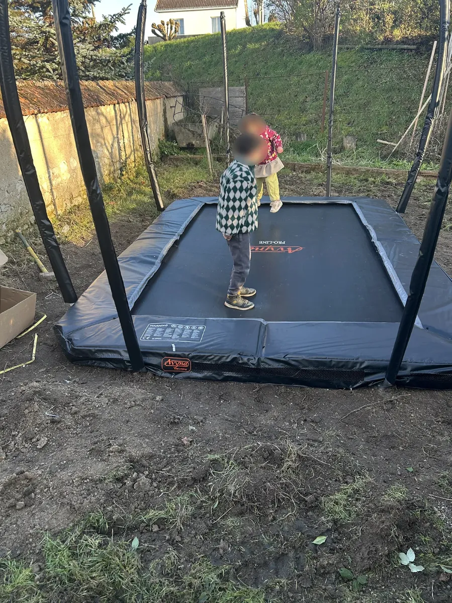 Trampoline enterré vu de côté dans un jardin en terre fraîchement remuée, enfants sur la toile