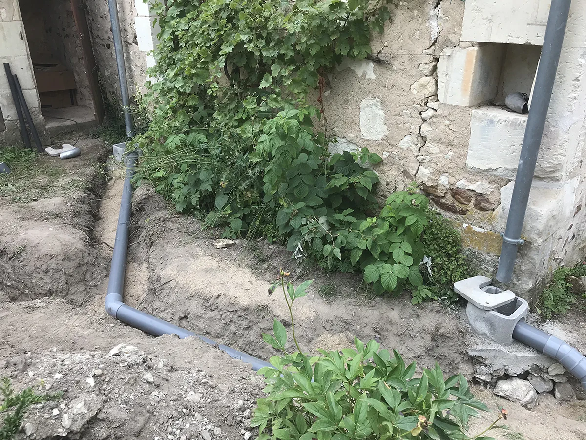 Descente d'eau pluviale en PVC raccordée à un regard en béton, au pied d'un mur de pierre ancien
